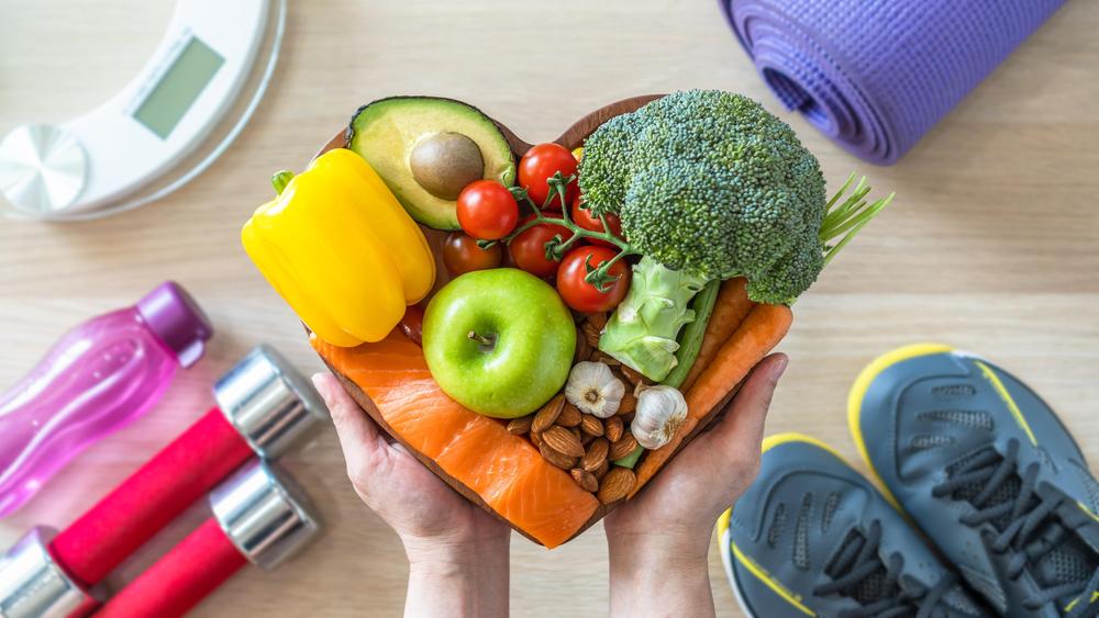 Im Vordergrund sind gesunde Nahrungsmittel abgebildet: Paprika, Avocado, Apfel, Tomaten, Brokkoli, Möhren, Knoblauch, Mandeln und Lachs. Im Hintergrund sieht man eine Waage, eine Yogamatte, Sportschuhe, Hanteln und eine Wasserflasche.
