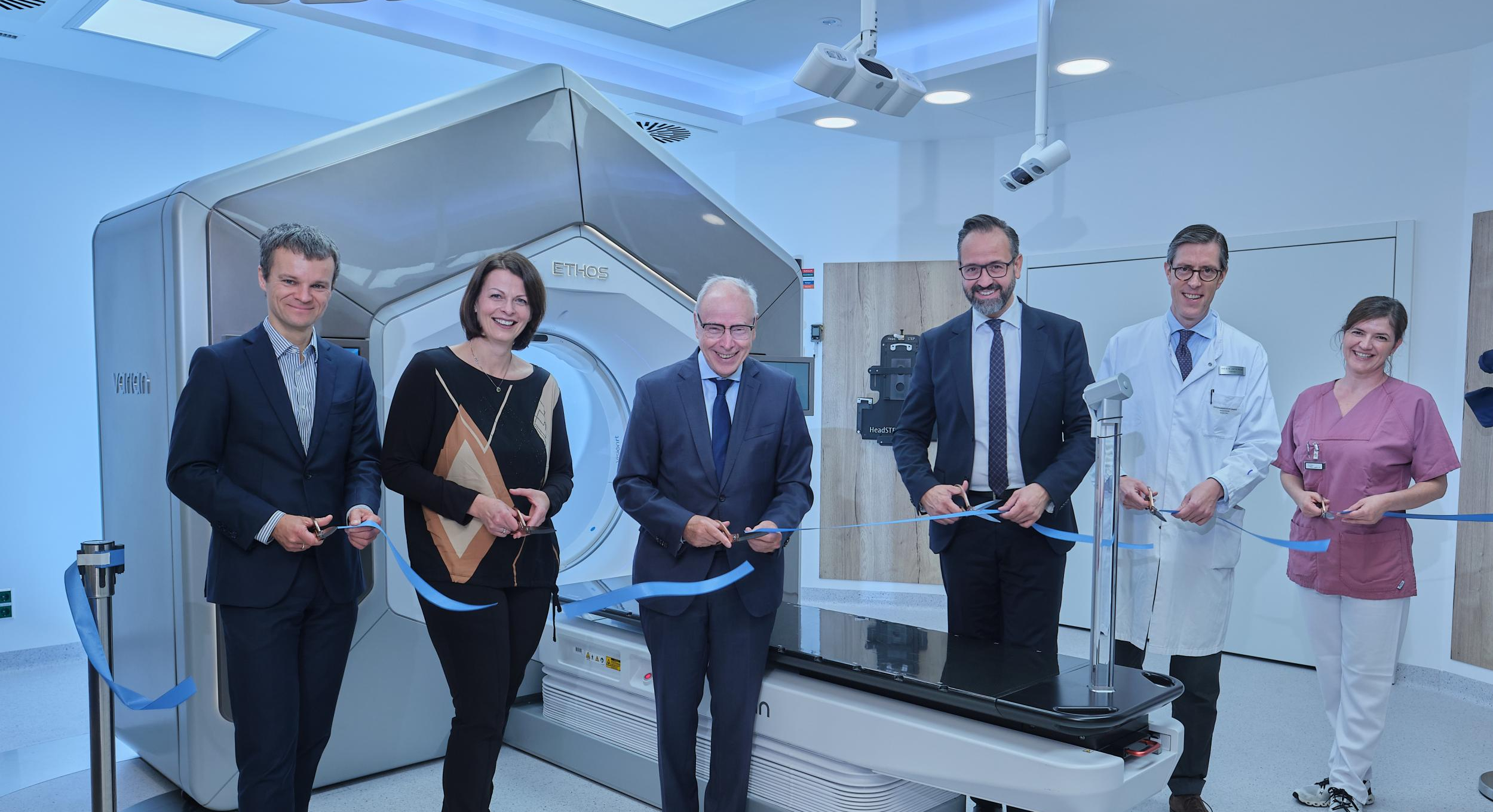 Im Hintergrund ist ein ETHOS-Linearbeschleuniger-Gerät zu sehen. Davor steht Sebastian Gemkow, Sächsischer Wissenschaftsminister, mit Vertreter:innen aus Politik, Wissenschaft und Gesundheitswesen, die das Gerät feierlich mit dem Durchschneiden eines Bandes eröffnen. 