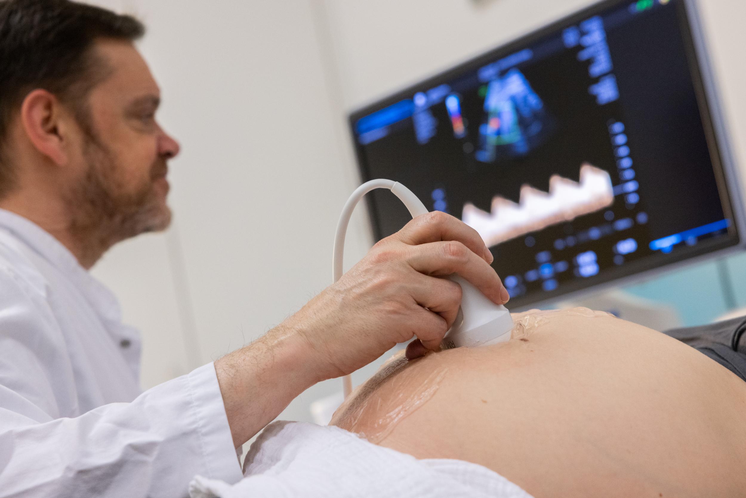 Ein Arzt hält ein Ultraschallgerät auf den Bauch einer Schwangeren. Im Hintergrund ist ein Bildschirm mit Ultraschallbildern zu sehen. 