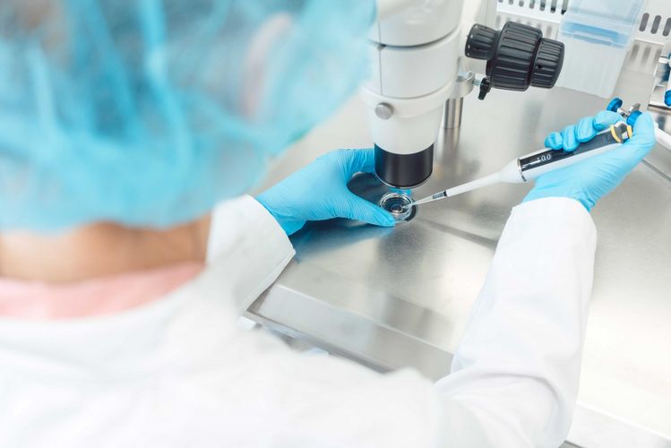 Das Bild zeugt eine Person mit steriler Kleidung und Handschuhen vor einem Mikroskop sitzen. In der Hand hält sie eine Pipette, die sie in eine Petrischale hält.