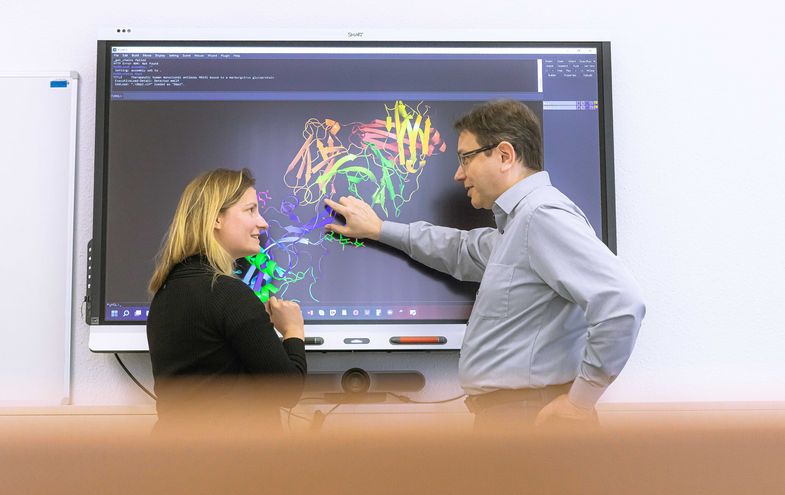 Zu sehen sind Prof. Dr. Jens Meiler und eine Frau, die vor einem großen Bildschirm stehen. Darauf sieht man ein Schema von Proteinen.