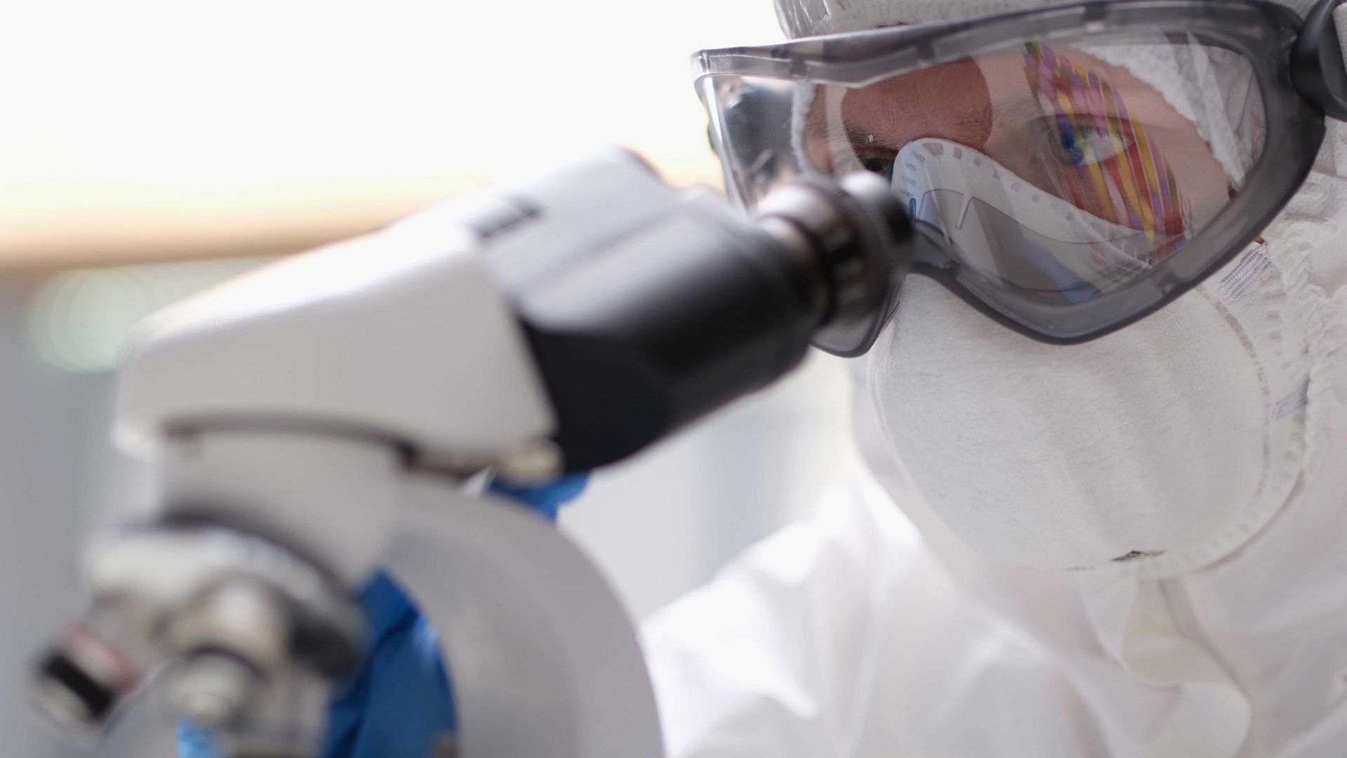 Auf dem Bild ist eine Person mit Schutzbrille und Schutzkleidung, die durch ein Mikroskop schaut.