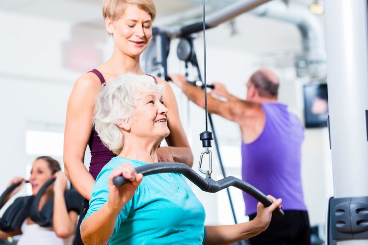 Zu sehen ist eine ältere Frau an einem Kabelzug in einem Fitnessstudio. Hinter ihr steht eine weitere Frau, die sie dabei beobachtet.