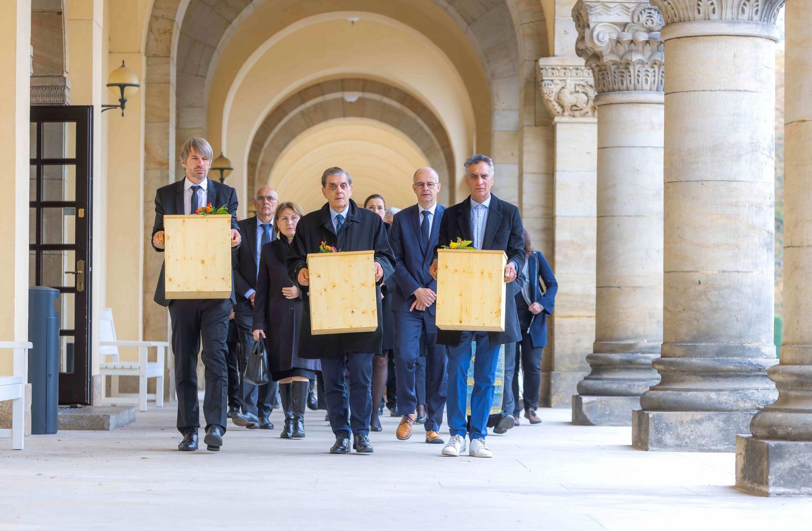 Man sieht einen Trauerzug mit drei symbolischen Särgen auf dem Weg zur Ruhestätte durch einen Bogengang. 