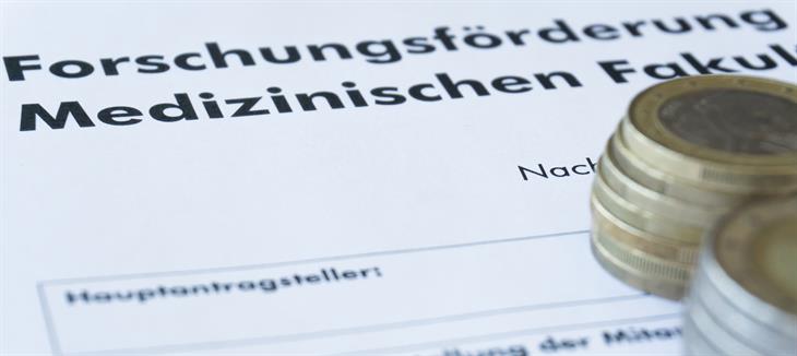 Man sieht ein Schriftstück mit dem Text „Forschungsförderung der Medizinischen Fakultät. Darauf befinden sich mehrere Euromünzen übereinander gestapelt. 