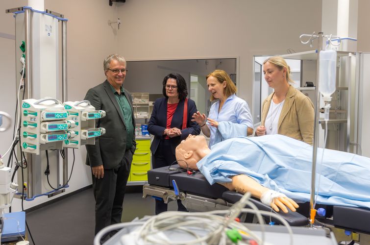 Vier Personen stehen vor einer Puppe auf einer Krankenliege in einem Krankenhausraum. Prof. Dr. Daisy Rotzoll redet. Links von ihr steht Prorektorin Prof. Dr. Brigitte Latzko. 