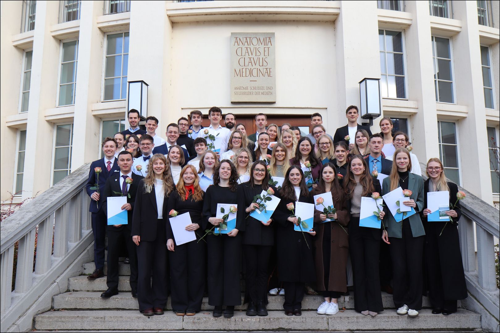 Großes Gruppenbild mit 44 Absolvent:innen in schwarzer eleganter Kleidung mit Urkunden und Rosen in der Hand auf den Treppenstufen vor einem Gebäude.  