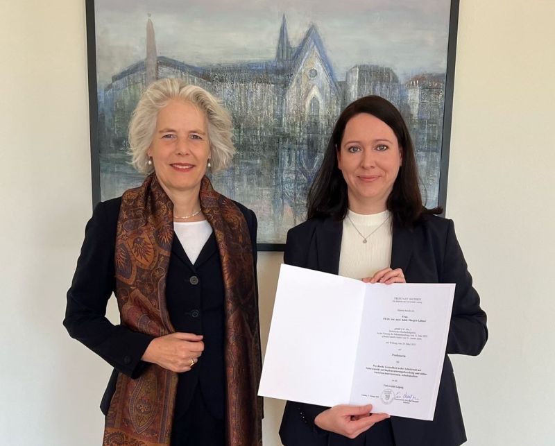 Zu sehen ist ein Bild von Prof. Margrit Löbner mit einer Urkunde in der Hand. Neben ihr steht eine andere Frau, die mit ihr in die Kamera lächelt.